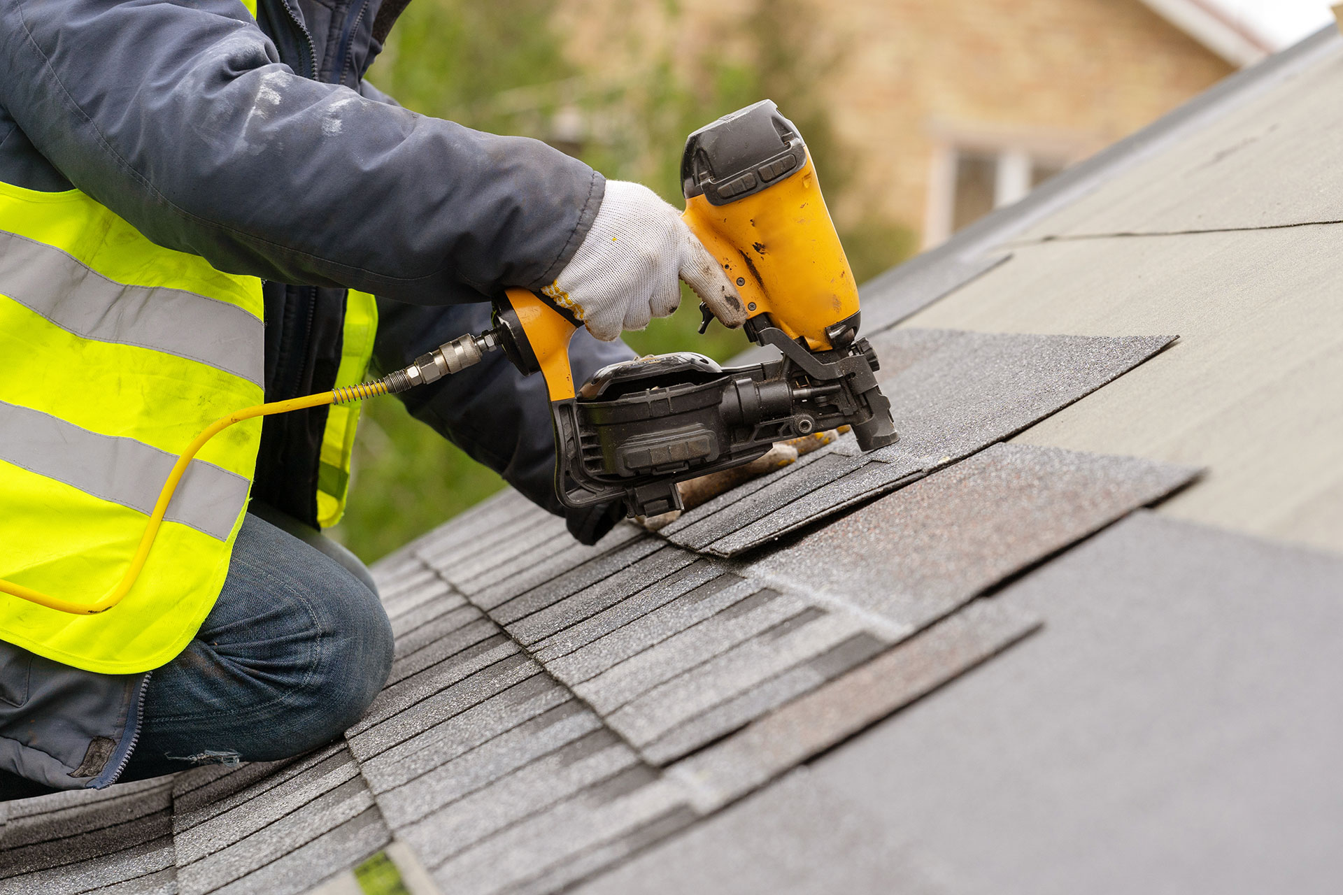 Roofer in worn denim jeans fastening new asphalt shingles with a nail gun on a Sacramento rooftop