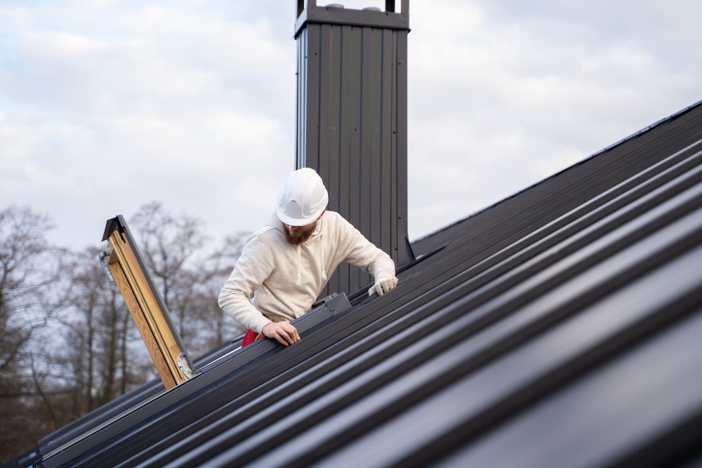 Roofer in a hard hat working on a dark standing-seam metal roof beside a skylight